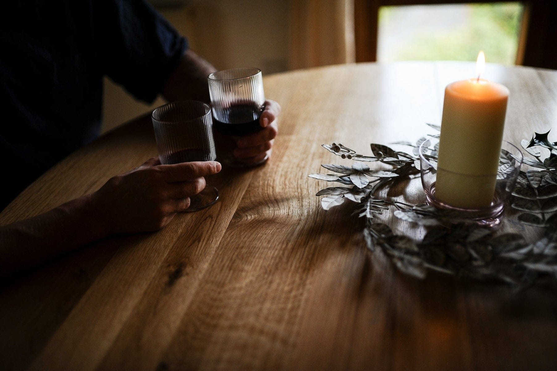 Person holding a glass of red wine at a wooden table with a lit candle and decorative elements.